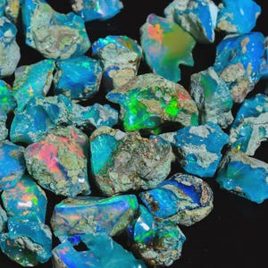 Può includere: Una collezione di gemme di opale grezze, che mostra una varietà di colori tra cui blu, verdi e lampi di rosso e arancione. Gli opali hanno una consistenza ruvida e naturale, con un mix di aree opache e traslucide. Le gemme sono sparse su una superficie scura.