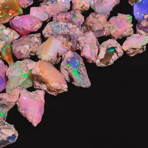 May include: A collection of raw opals in various colors, including pink, orange, blue, and green. The opals have a rough, natural texture and are set against a black background. The gemstones display a play of color, with flashes of iridescence.