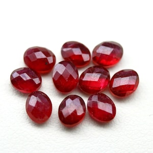 May include: A collection of nine oval-shaped, faceted red gemstones arranged in a cluster. Each stone features a polished surface with geometric facets that reflect light, showcasing a deep, vibrant red color throughout the entire set of loose gems.