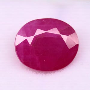May include: A faceted, oval-shaped ruby gemstone. The gem is a deep, saturated red color with a polished surface. The facets create a pattern of light and shadow across the surface of the stone.