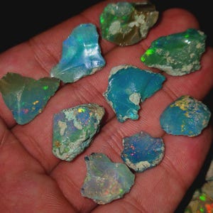 May include: A collection of rough opal gemstones in various shapes and sizes, held in a hand. The opals display a range of colors, including blues, greens, and flashes of other hues. The stones have a natural, unpolished appearance.