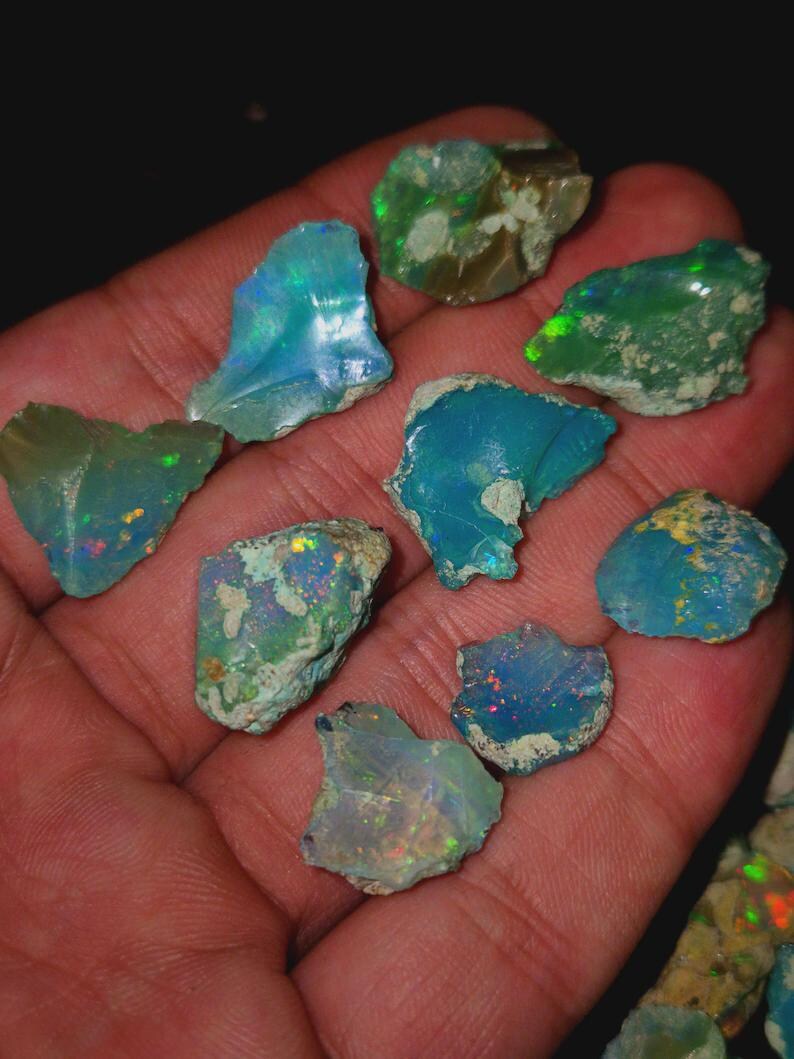 May include: A collection of rough opal gemstones in various shapes and sizes, held in a hand. The opals display a range of colors, including blues, greens, and flashes of other hues. The stones have a natural, unpolished appearance.