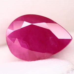 May include: A faceted, pear-shaped ruby gemstone. The gem is a deep, saturated pinkish-red color with multiple facets that reflect light. The gemstone is set against a white background.