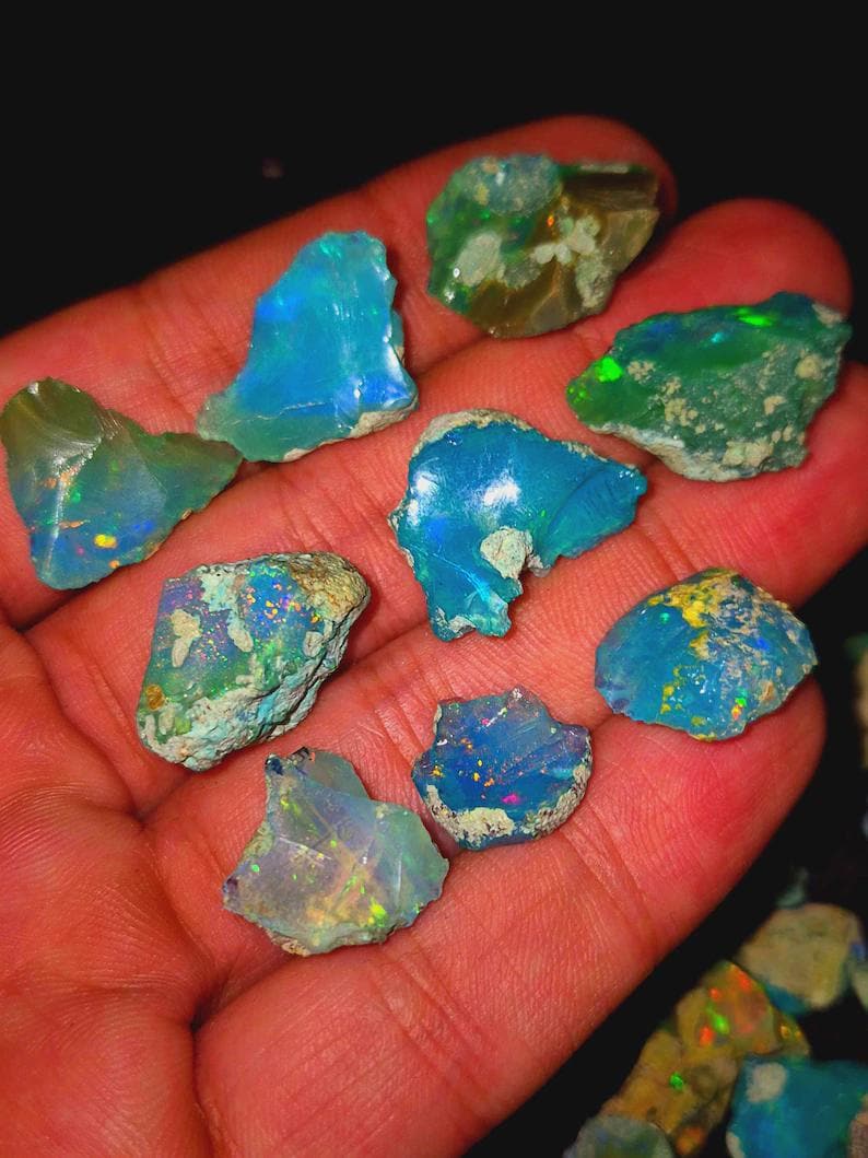 May include: A collection of raw opal gemstones in various shapes and sizes. The opals display a vibrant play-of-color, with flashes of blue, green, and yellow. The gemstones are held in a hand, showcasing their natural beauty and unique patterns.