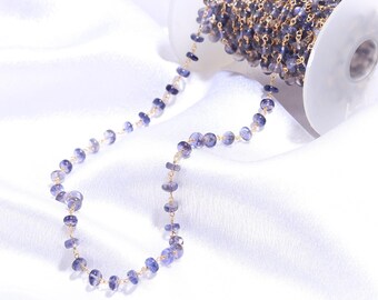 May include: A spool of gold-toned wire chain featuring small, faceted, translucent purple iolite beads spaced evenly along the length. The chain is wound around a white plastic spool, with a portion of the beaded chain extending outward.