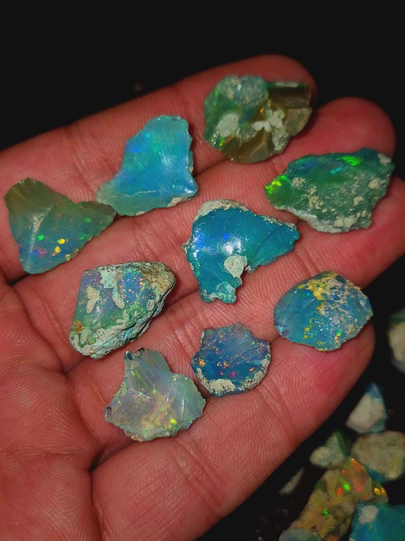 May include: A collection of raw opal gemstones in various shapes and sizes. The opals display a range of colors, including blues, greens, and flashes of iridescent hues. The rough texture of the stones is visible, showcasing their natural state.