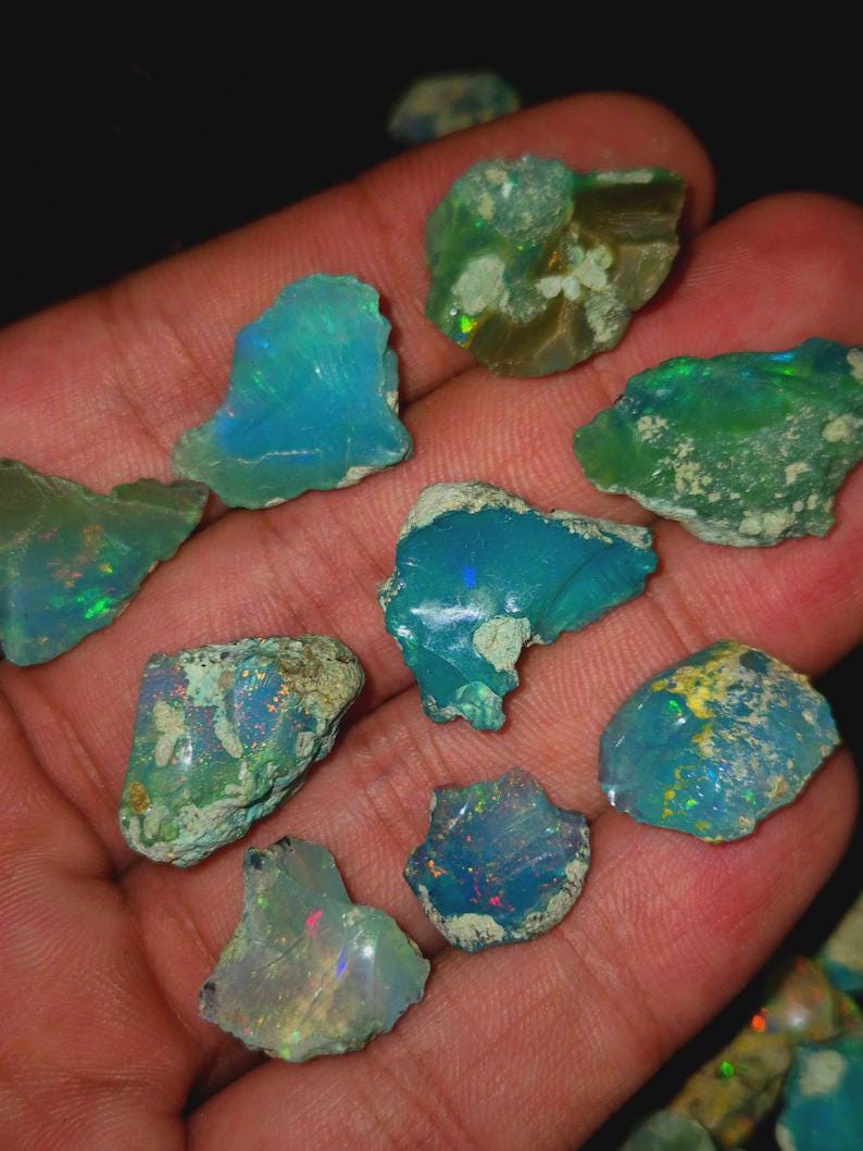 May include: A collection of raw opal gemstones in various shapes and sizes. The opals display a range of colors, including blues, greens, and flashes of iridescent hues. The rough texture of the stones is visible, showcasing their natural state.