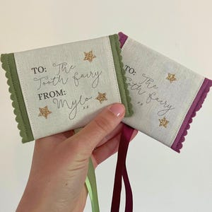 May include: Two small fabric tooth fairy pouches. One is green with a scalloped edge, the other is purple. Both have the text "TO: The Tooth Fairy FROM: Mylo" and gold glitter star accents.