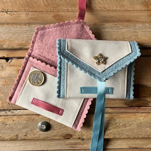 May include: Handmade fabric wallets in a light color with scalloped edges and a star embellishment. One wallet is open, revealing a coin. The wallets have pink, blue, and light blue accents, with a ribbon closure. The background is a wooden surface.