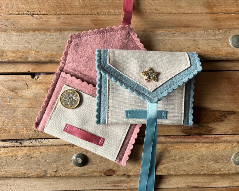 May include: Handmade fabric wallets in a light color with scalloped edges and a star embellishment. One wallet is open, revealing a coin. The wallets have pink, blue, and light blue accents, with a ribbon closure. The background is a wooden surface.
