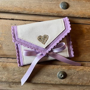 May include: A small, off-white fabric wallet with a lavender scalloped trim and a sparkling heart embellishment. A lavender ribbon is tied around the wallet. The wallet is resting on a wooden surface.