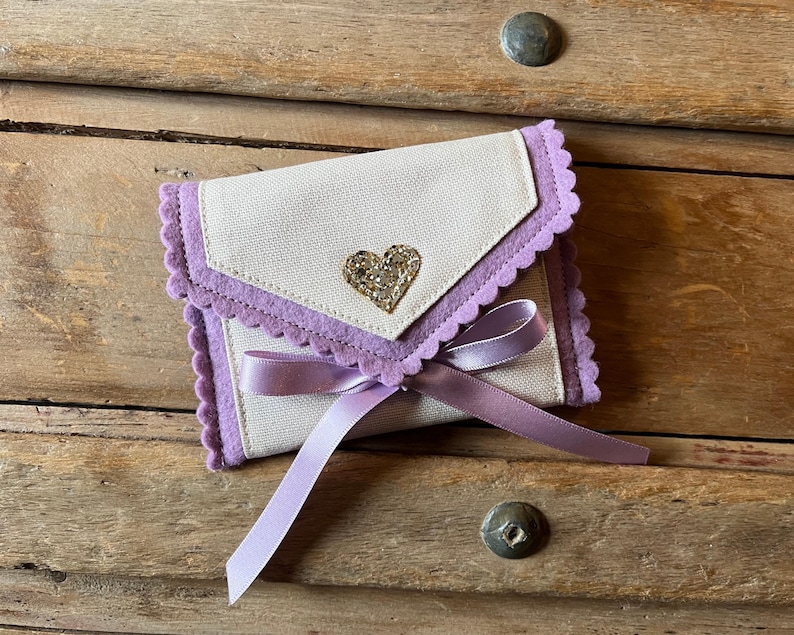 May include: A small, off-white fabric wallet with a lavender scalloped trim and a sparkling heart embellishment. A lavender ribbon is tied around the wallet. The wallet is resting on a wooden surface.