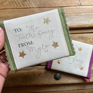 May include: Two small, rectangular tooth fairy pouches. One is held in a hand. The pouches are off-white with green and purple trim. Text on the pouches reads "TO: The Tooth Fairy FROM: Mylo" with gold star accents.