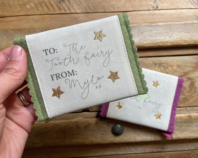 May include: Two small, rectangular tooth fairy pouches. One is held in a hand. The pouches are off-white with green and purple trim. Text on the pouches reads "TO: The Tooth Fairy FROM: Mylo" with gold star accents.
