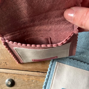 May include: A small, open, dusty rose felt pouch with a scalloped edge and a light beige rectangular patch. The pouch is held open, revealing a small pocket inside. A second, similar pouch in light blue is visible in the background.