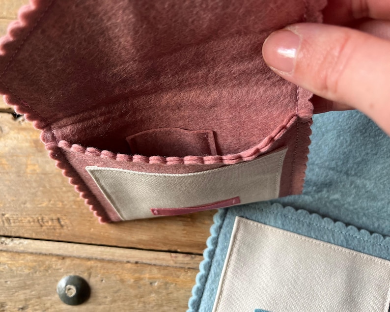 May include: A small, open, dusty rose felt pouch with a scalloped edge and a light beige rectangular patch. The pouch is held open, revealing a small pocket inside. A second, similar pouch in light blue is visible in the background.