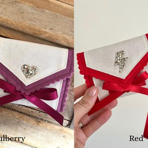 May include: Two small, decorative fabric pouches. One is mulberry with a heart-shaped jewel and a burgundy ribbon. The other is red with a lightning bolt jewel and a red ribbon. The pouches have a scalloped edge.