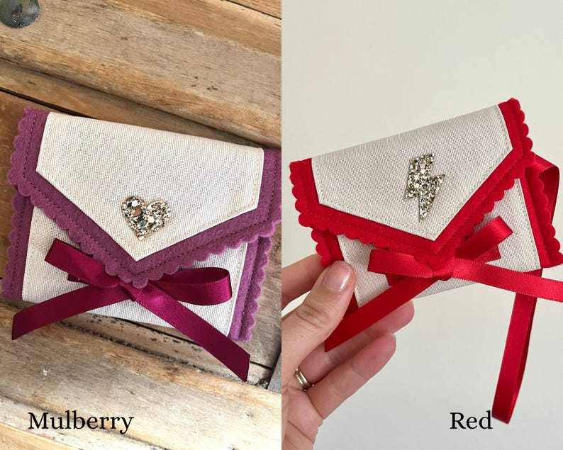 May include: Two small, decorative fabric pouches. One is mulberry with a heart-shaped jewel and a burgundy ribbon. The other is red with a lightning bolt jewel and a red ribbon. The pouches have a scalloped edge.
