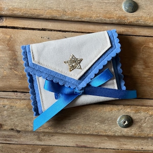 May include: A small, handmade envelope-style pouch with a light beige fabric body and blue felt trim. A gold glitter star embellishes the front flap, and a blue satin ribbon secures the pouch. The pouch is set against a wooden background.