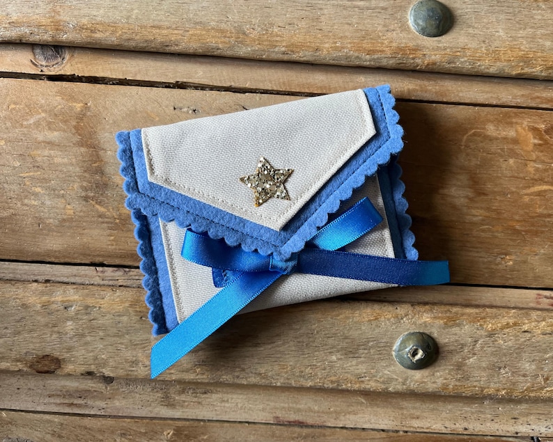 May include: A small, handmade envelope-style pouch with a light beige fabric body and blue felt trim. A gold glitter star embellishes the front flap, and a blue satin ribbon secures the pouch. The pouch is set against a wooden background.