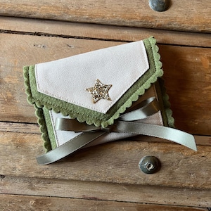 May include: A small, handmade pouch with a star embellishment. The pouch features a light beige fabric with a scalloped green trim and a green ribbon. A gold star is centered on the front. The pouch is closed with a green ribbon.