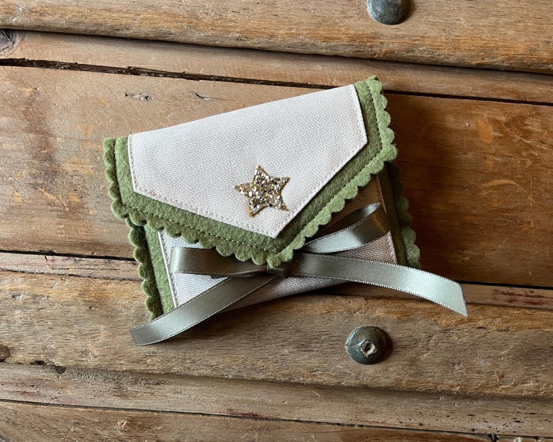 May include: A small, handmade pouch with a star embellishment. The pouch features a light beige fabric with a scalloped green trim and a green ribbon. A gold star is centered on the front. The pouch is closed with a green ribbon.