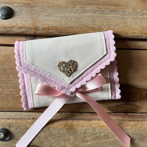 May include: A small, off-white fabric envelope-style pouch with a pink scalloped trim and a gold glitter heart. A pink satin ribbon is tied in a bow at the bottom. The pouch is closed.