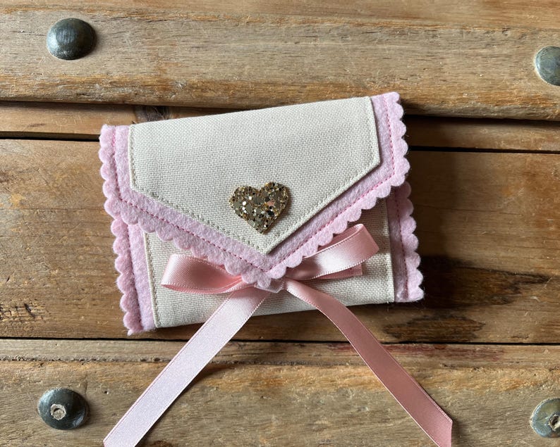 May include: A small, off-white fabric envelope-style pouch with a pink scalloped trim and a gold glitter heart. A pink satin ribbon is tied in a bow at the bottom. The pouch is closed.