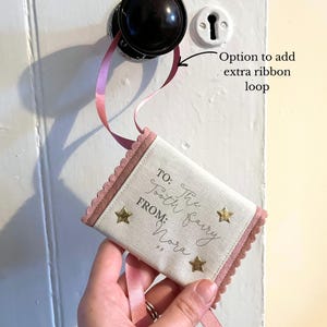 May include: A small, rectangular tooth fairy pillow with a pink scalloped border and ribbon. The pillow is cream-colored with gold star accents and text that reads "TO: The Tooth Fairy FROM: Nora". The pillow is hanging from a doorknob.