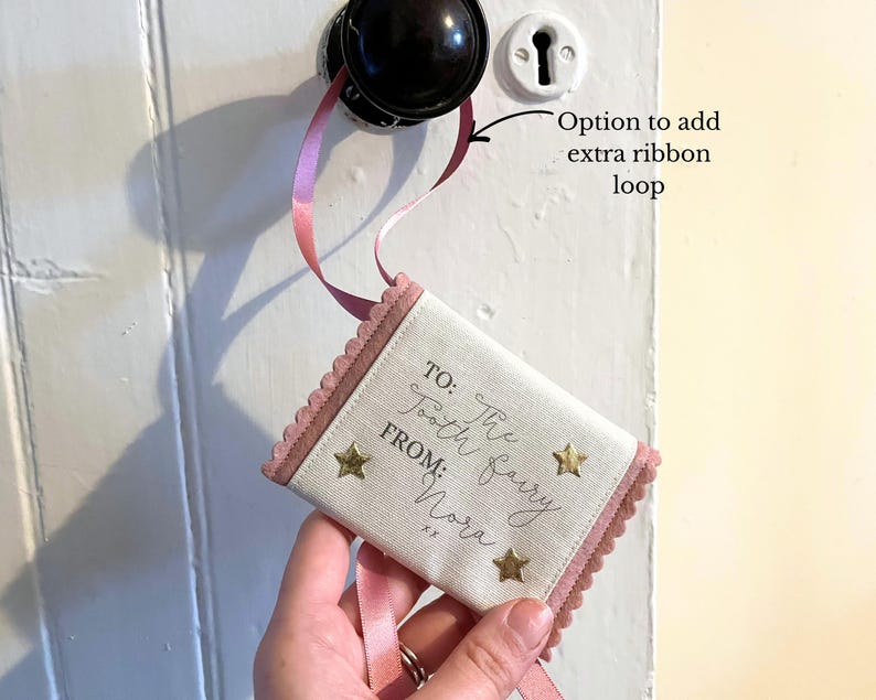 May include: A small, rectangular tooth fairy pillow with a pink scalloped border and ribbon. The pillow is cream-colored with gold star accents and text that reads "TO: The Tooth Fairy FROM: Nora". The pillow is hanging from a doorknob.