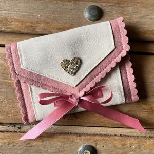May include: A small, cream-colored fabric envelope with a pink scalloped trim and a sparkling heart-shaped embellishment. A pink satin ribbon is tied in a bow at the bottom. The envelope is resting on a wooden surface.