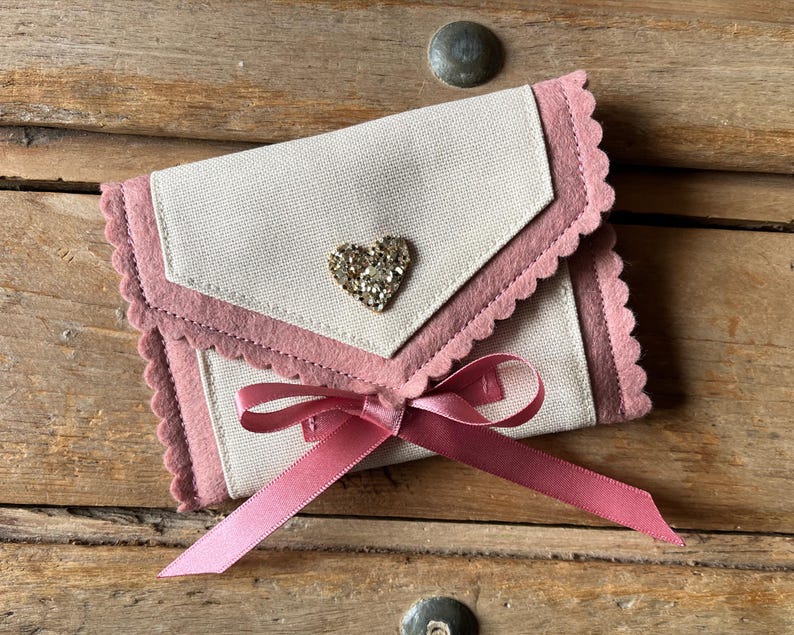 May include: A small, cream-colored fabric envelope with a pink scalloped trim and a sparkling heart-shaped embellishment. A pink satin ribbon is tied in a bow at the bottom. The envelope is resting on a wooden surface.