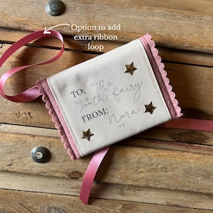 May include: A small, rectangular tooth fairy pouch with a pink ribbon and scalloped edges. The pouch is cream-colored with gold star accents and text that reads "TO: The Tooth Fairy FROM: Nora".