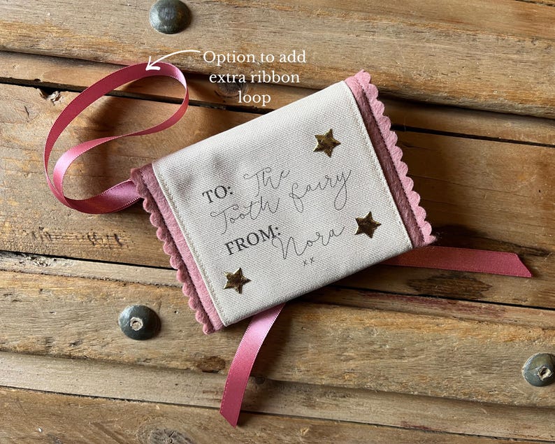 May include: A small, rectangular tooth fairy pouch with a pink ribbon and scalloped edges. The pouch is cream-colored with gold star accents and text that reads "TO: The Tooth Fairy FROM: Nora".