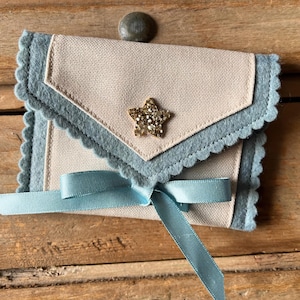 May include: A small, handmade envelope-style pouch with a light blue scalloped edge and a light tan center. A gold glitter star is affixed to the front. A light blue ribbon is tied in a bow at the bottom. The pouch is on a wooden surface.