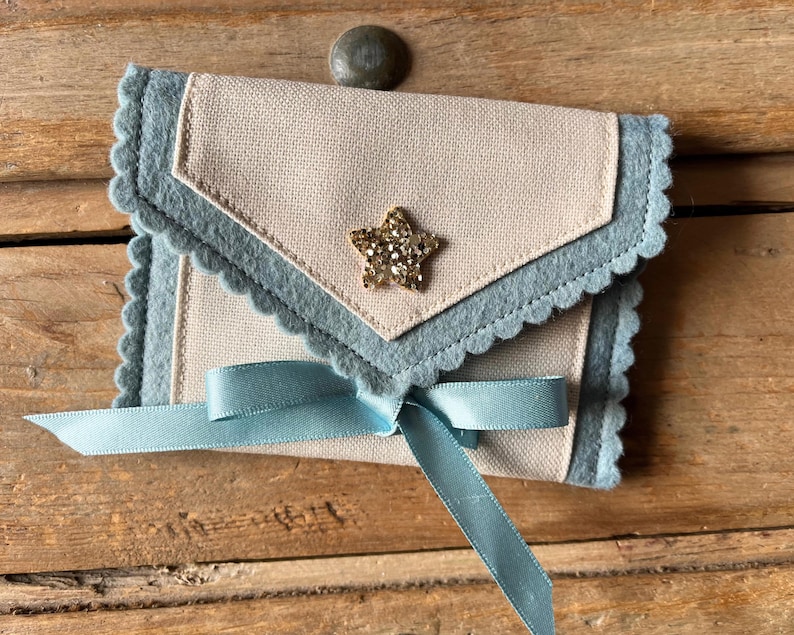 May include: A small, handmade envelope-style pouch with a light blue scalloped edge and a light tan center. A gold glitter star is affixed to the front. A light blue ribbon is tied in a bow at the bottom. The pouch is on a wooden surface.