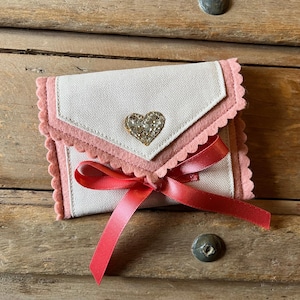 May include: A small, handmade wallet with a light-colored fabric exterior and pink felt trim. A sparkly heart embellishment is centered on the flap. A red ribbon is tied in a bow. The wallet is on a wooden surface.