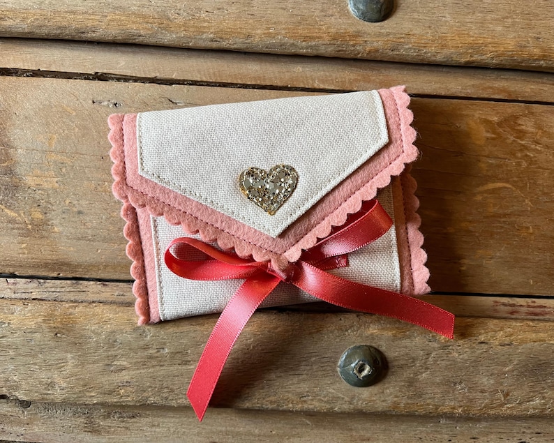 May include: A small, handmade wallet with a light-colored fabric exterior and pink felt trim. A sparkly heart embellishment is centered on the flap. A red ribbon is tied in a bow. The wallet is on a wooden surface.