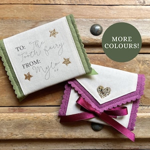 May include: Two small, fabric tooth fairy pouches. One is white with green trim and gold stars, the text reads "TO: The Tooth fairy FROM: Mylo". The other is white with purple trim, a heart embellishment, and a purple ribbon.