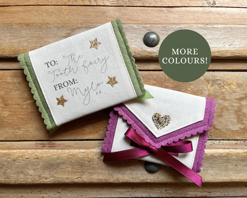 May include: Two small, fabric tooth fairy pouches. One is white with green trim and gold stars, the text reads "TO: The Tooth fairy FROM: Mylo". The other is white with purple trim, a heart embellishment, and a purple ribbon.