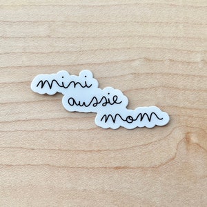 May include: White sticker with black text that reads "mini aussie mom" on a light wood background.