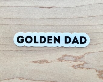 Könnte beinhalten: Weißer Aufkleber mit schwarzem Text "GOLDEN DAD" auf hellem Holzgrund.