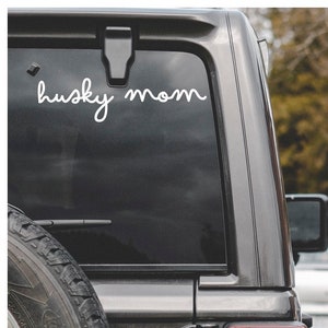 May include: A black SUV rear window with a white cursive decal that reads "husky mom." The vehicle has a spare tire, a red taillight, and a black door handle.