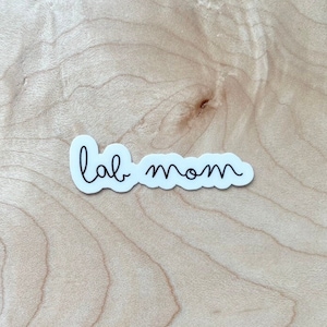 May include: White sticker with black text that reads "lab mom" on a light brown wood background.