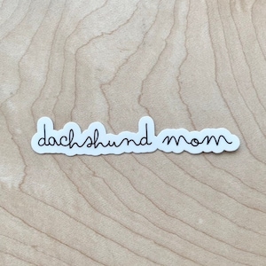 May include: White sticker with black cursive text that reads "dachshund mom" on a light brown wood background.