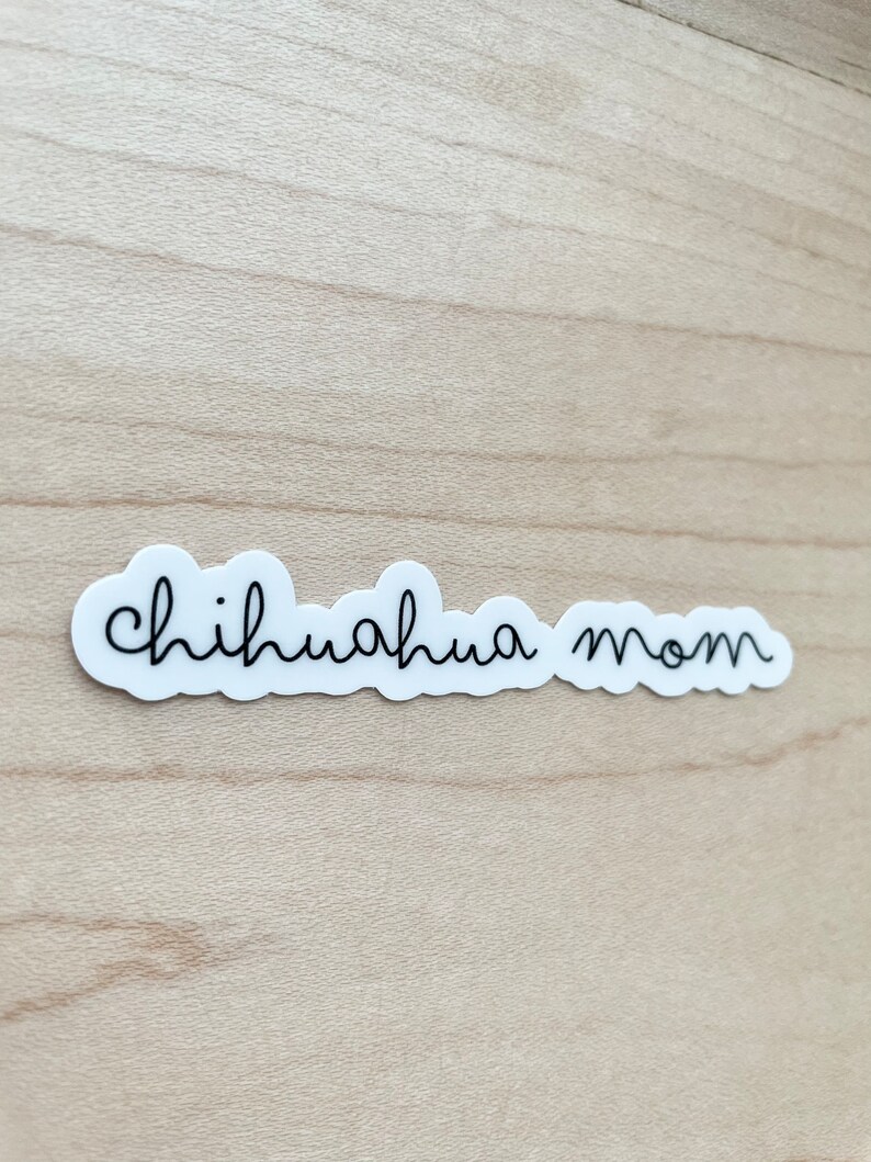 May include: White sticker with black text that reads "chihuahua mom" on a light brown wooden surface.