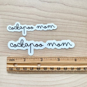 May include: Two white stickers with black text that reads "cockapoo mom" on a wooden surface with a ruler for scale.
