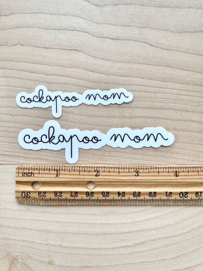 May include: Two white stickers with black text that reads "cockapoo mom" on a wooden surface with a ruler for scale.