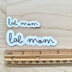 May include: Two white stickers with black text that reads "lab mom". The stickers are on a wooden surface with a ruler underneath.