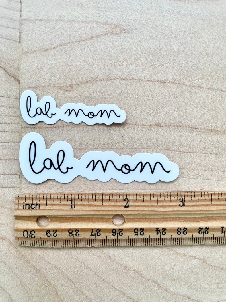 May include: Two white stickers with black text that reads "lab mom". The stickers are on a wooden surface with a ruler underneath.